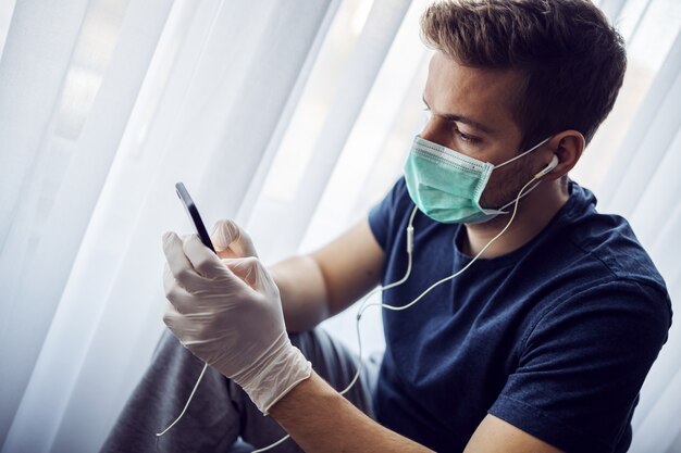 Hombre con máscara y guantes escuchando música y escribiendo en el teléfono inteligente. Quédate en casa. Pandemia mundial, coronavirus.