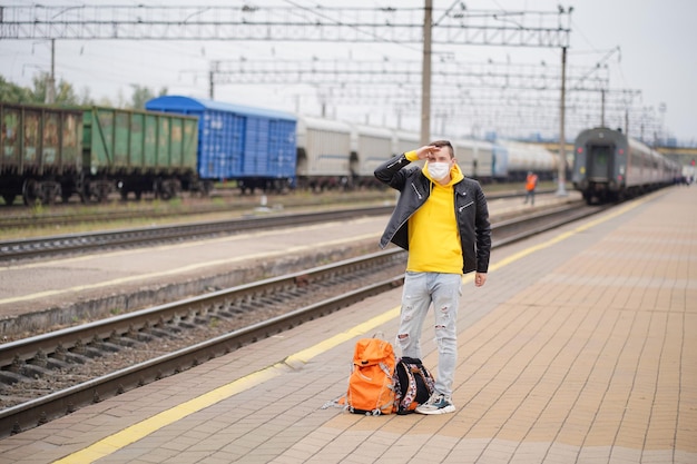 Hombre con máscara médica se para en la plataforma esperando el tren Pasajero masculino con máscara protectora con mochilas busca el tren en la plataforma del ferrocarril durante la pandemia de coronavirus