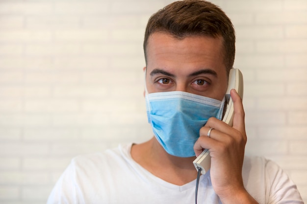 Un hombre con una máscara médica de protección facial para protegerse del virus, dentro de su casa, hablando por teléfono.