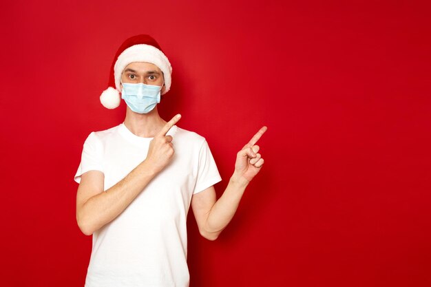 Hombre en máscara médica con Santa Claus Christmas hat point asiento vacío aislado fondo de estudio rojo