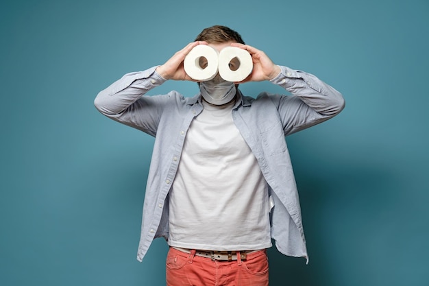 El hombre con una máscara médica sostiene dos rollos de papel higiénico en las manos y los mira como binoculares