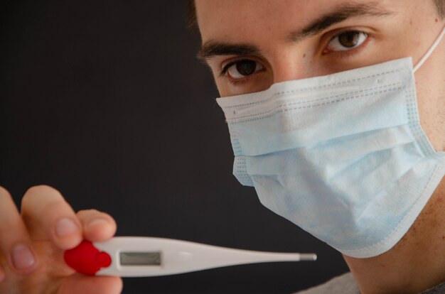 Foto un hombre con una máscara médica sostiene, mira un termómetro.