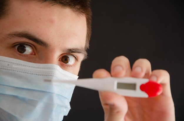 Un hombre con una máscara médica sostiene, mira un termómetro.