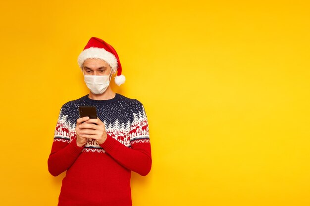 Hombre con máscara médica suéter de Navidad gorro de Papá Noel tiene smartphone en sus manos escribe un mensaje