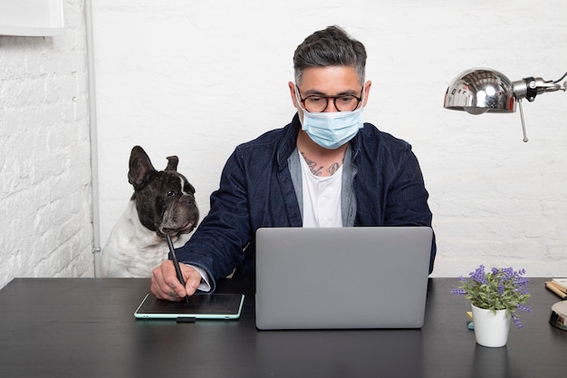 Hombre en máscara médica trabajando en un diseño creativo desde casa con su perro sentados juntos en el espacio de trabajo.