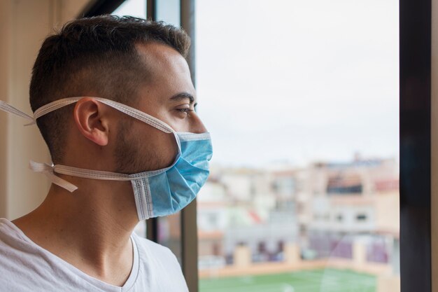 Hombre con una máscara medicinal para protegerse contra COVID-19, confinado en su hogar, mirando desconsolado por la ventana.