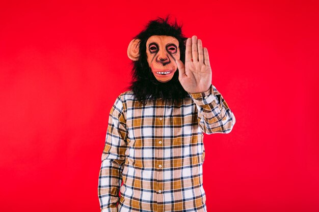 Hombre con máscara de mono chimpancé y camisa a cuadros haciendo la señal de alto con la mano en el fondo rojo