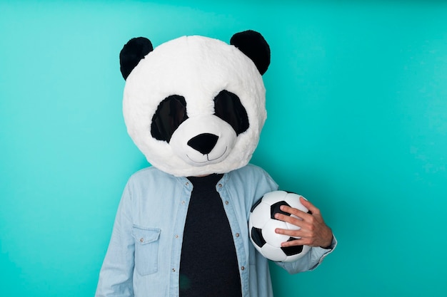 Hombre con máscara de panda sosteniendo una pelota de fútbol sobre pared azul