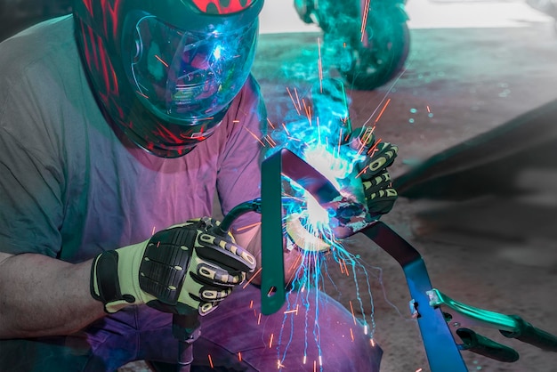 Un hombre con una máscara protectora trabaja con una herramienta de soldadura para metal la reconstrucción de un producto metálico usando soldadura la profesión de soldadorel proceso de soldadura metalchispas y humo durante el metal