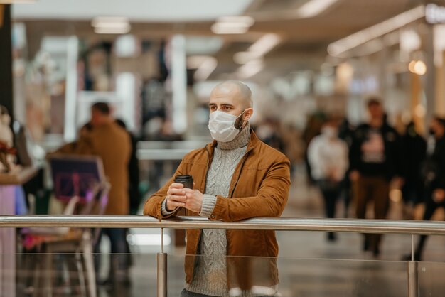 Foto un hombre con mascarilla para evitar la propagación del coronavirus sostiene una taza de café mientras espera en el centro comercial. un tipo calvo con una mascarilla quirúrgica mantiene la distancia social.