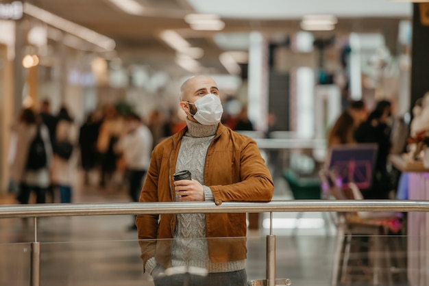Foto un hombre con mascarilla para evitar la propagación del coronavirus sostiene una taza de café mientras espera en el centro comercial. un tipo calvo con una mascarilla quirúrgica mantiene la distancia social.