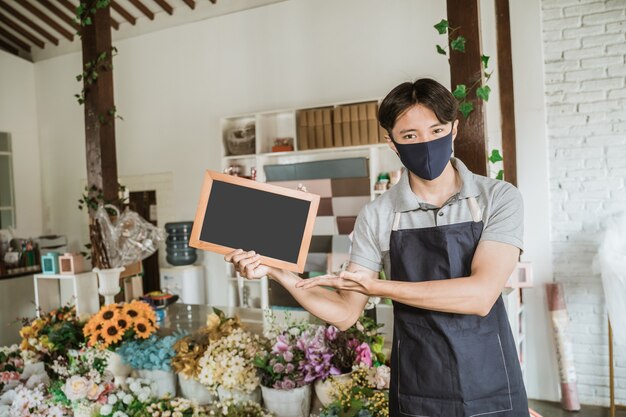 Hombre con mascarilla floristería de pie sosteniendo pequeñas flores frescas de pizarra en blanco en su tienda de flores siguiendo el protocolo saludable