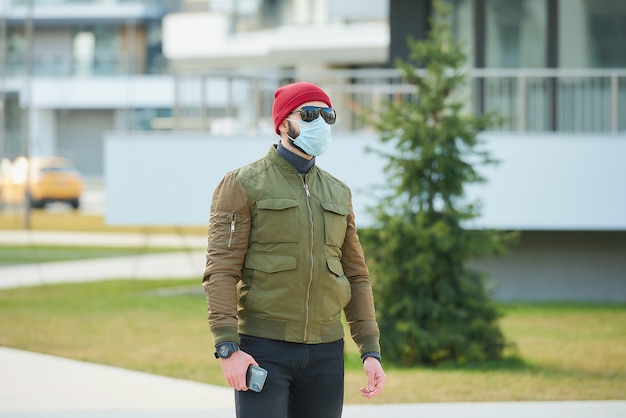 Un hombre con una mascarilla médica para evitar la propagación del coronavirus sosteniendo su teléfono inteligente en una calle acogedora.