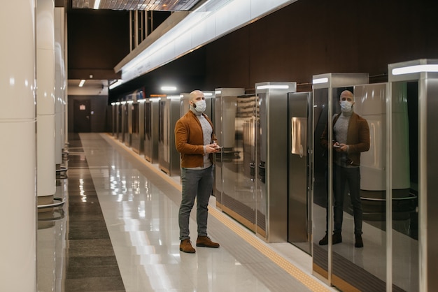 Un hombre con una mascarilla médica para evitar la propagación del coronavirus sostiene un teléfono inteligente mientras espera un tren en el metro. Un tipo calvo con una mascarilla quirúrgica mantiene la distancia social.