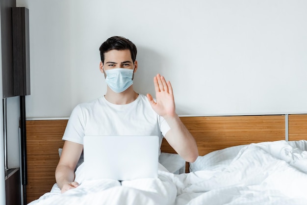 Hombre con mascarilla médica saludando con la mano a la cámara mientras está sentado con una laptop en la cama