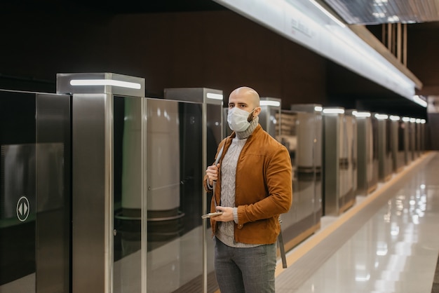 Un hombre con una mascarilla médica sostiene un teléfono inteligente y mira hacia un lado mientras espera un tren en la plataforma del metro. Un tipo calvo con una mascarilla quirúrgica mantiene la distancia social.