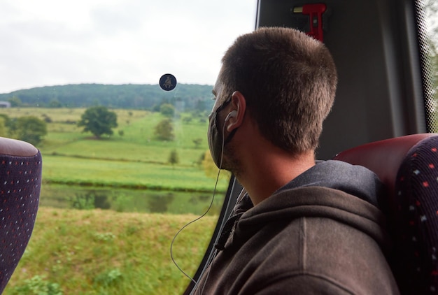 Un hombre con una mascarilla negra sentado en el transporte público y usando audífonos durante el covid-19