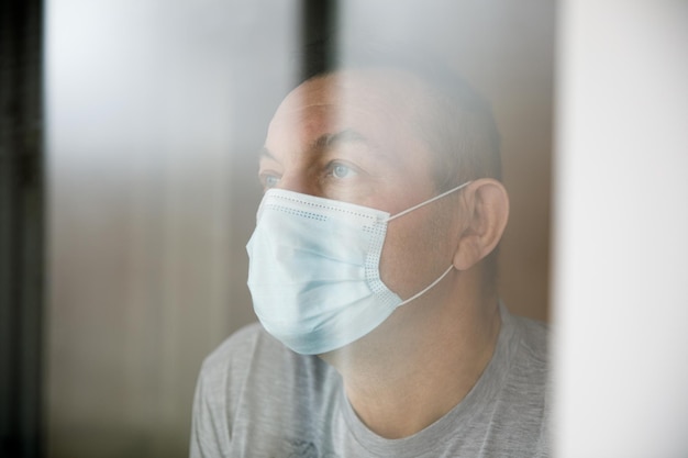 Hombre con una mascarilla sentado en una casa de cuarentena y mirando por la ventana.