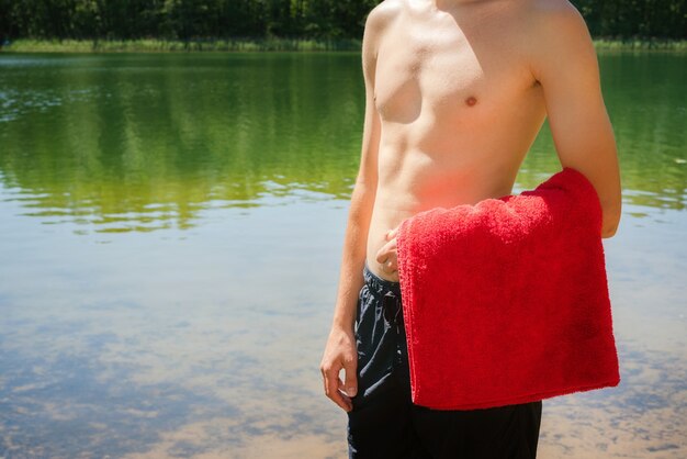 Hombre masculino atlético joven y en forma de pie en la orilla del lago sin una camisa mientras sostiene una toalla roja.