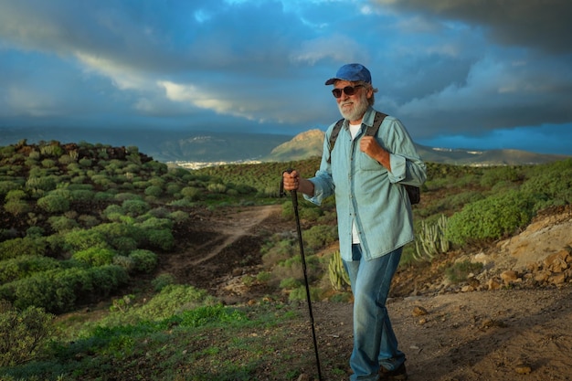 Hombre mayor activo al aire libre disfrutando del senderismo a la luz del atardecer Anciano caucásico con sombrero y mochila caminando con bastón en el paisaje tropical de montaña Cielo dramático de montaña verde