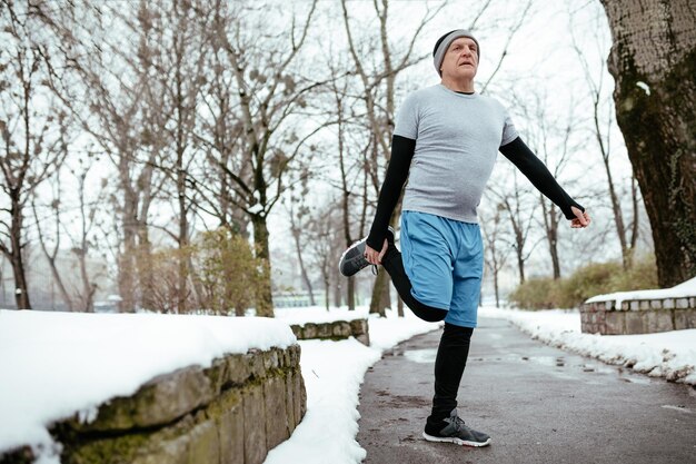 Hombre mayor activo estirándose y haciendo ejercicios antes de trotar, en un parque público durante el entrenamiento de invierno afuera. Copie el espacio.