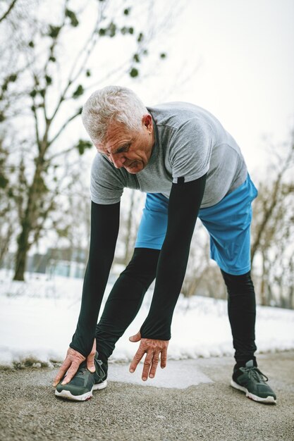 Hombre mayor activo estirándose y haciendo ejercicios en un parque público durante el entrenamiento de invierno afuera.