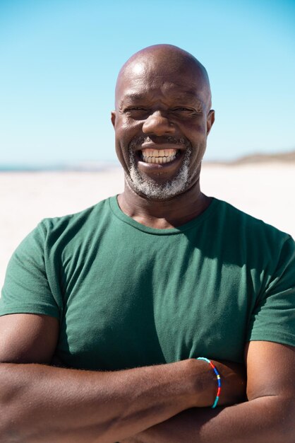 Hombre mayor afroamericano calvo y despreocupado con los brazos cruzados sonriendo en la playa contra el cielo despejado. Copie el espacio, el verano, el retrato, el estilo de vida inalterado, las vacaciones, la jubilación, el disfrute y el concepto de naturaleza.