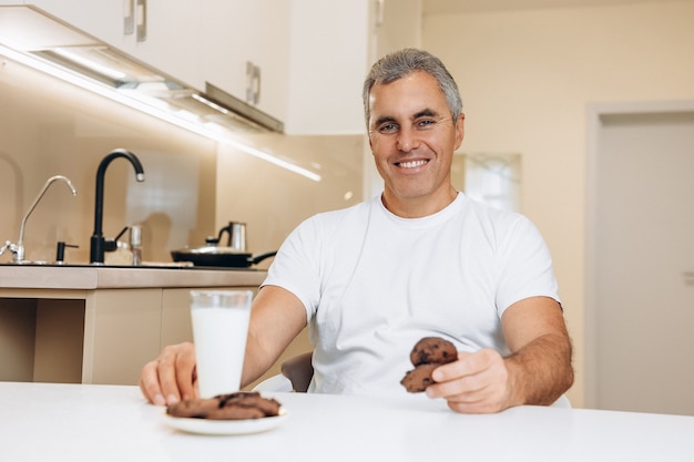 Hombre mayor alegre en camiseta blanca tiene un vaso de galletas de chocolate y leche