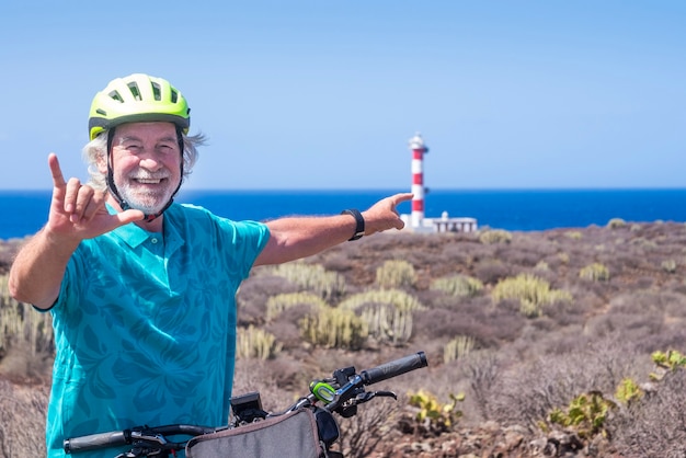 Hombre mayor alegre con casco amarillo disfrutando de una excursión con bicicleta cerca de un faro