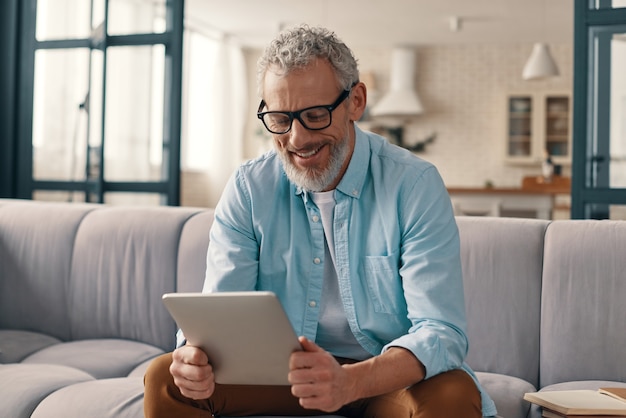 Hombre mayor alegre en ropa casual con tableta digital mientras está sentado en el sofá en casa