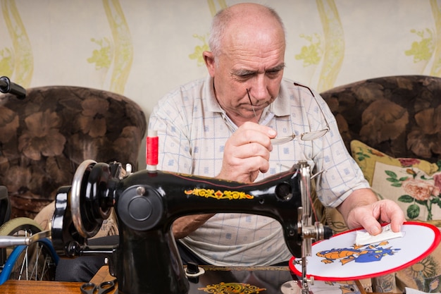 Hombre mayor con anteojos inspeccionando el trabajo de la punta de la aguja en colgar en la pared usando la antigua máquina de coser manual en casa en la sala de estar