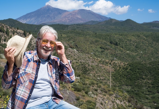 Foto hombre mayor atractivo con barba blanca disfrutando de una excursión al aire libre en el paisaje de montaña, sonriendo mirando a cámara. volcán del teide al fondo.