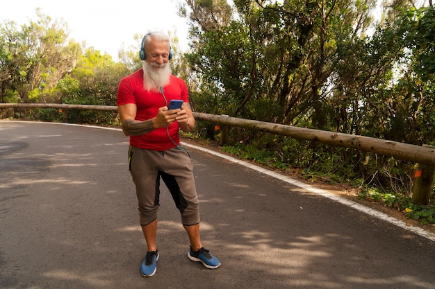 Foto hombre mayor con barba blanca corriendo al aire libre en la naturaleza sosteniendo el teléfono y eligiendo música