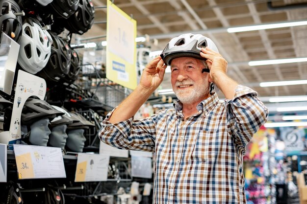 Hombre mayor con barba en camisa selecciona un casco protector de bicicleta en una tienda de deportes Sostiene un casco blanco mientras lo prueba en su cabeza Concepto de consumismo