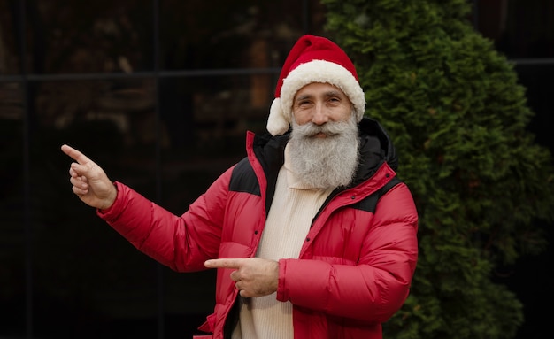 Hombre mayor con barba en el sombrero rojo de santa que presenta algo al aire libre. Navidad. Año nuevo.