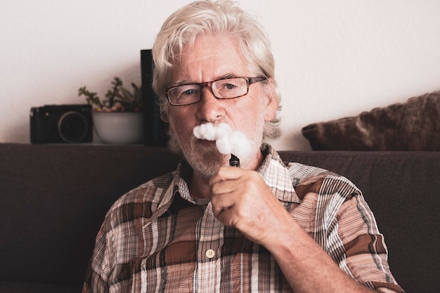 Hombre mayor barbudo fumando cigarrillo electrónico sentado en el sofá en casa - nueva tecnología para un humo más saludable