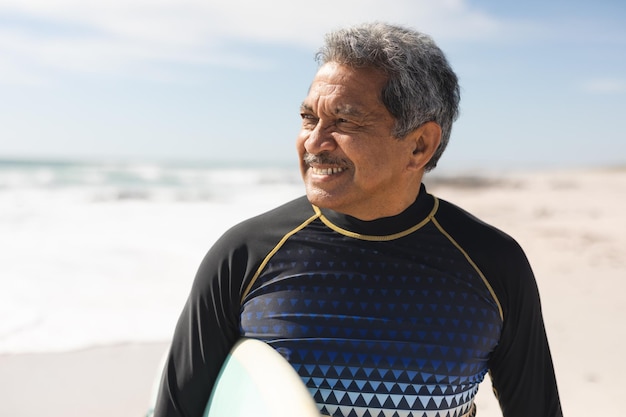 Un hombre mayor birracial jubilado sonriente mirando hacia otro lado mientras estaba de pie con una tabla de surf en la playa en un día soleado