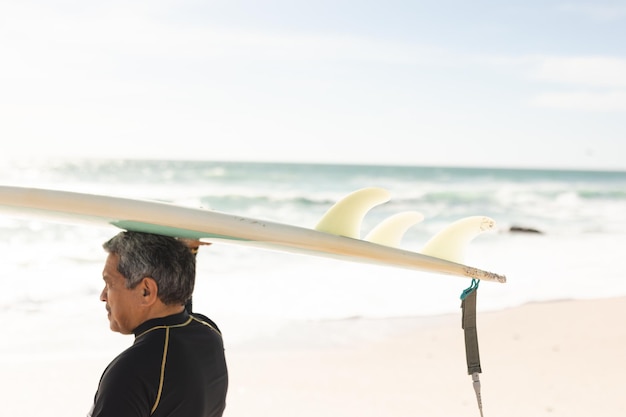 Hombre mayor birracial llevando tablas de surf sobre la cabeza en la playa contra el cielo durante el día soleado