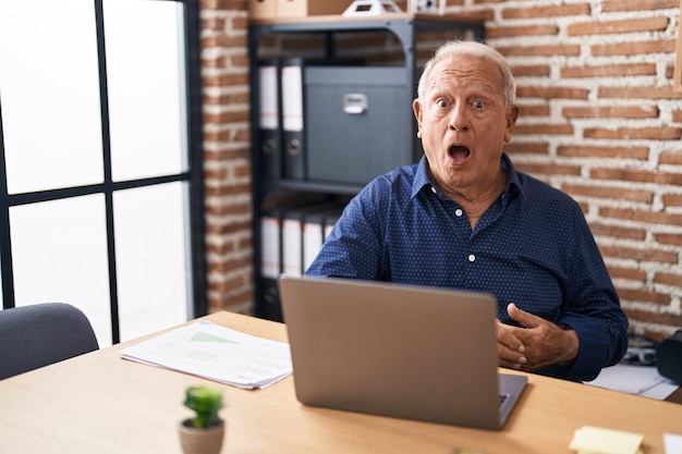 Hombre mayor con cabello gris trabajando usando computadora portátil en la oficina asustado y asombrado con la boca abierta por cara de incredulidad sorpresa