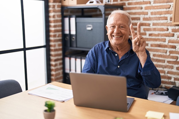 Hombre mayor con cabello gris trabajando usando una computadora portátil en la oficina sonriendo con cara feliz guiñando el ojo a la cámara haciendo el signo de victoria número dos