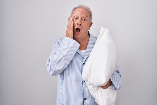 Hombre mayor con cabello gris usando pijama abrazando almohada asustado y conmocionado, sorpresa y expresión asombrada con las manos en la cara