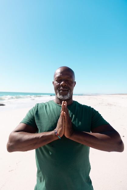 Hombre mayor calvo afroamericano con los ojos cerrados meditando en posición de oración en la playa, espacio para copiar. Verano, cielo, inalterado, ejercicio, yoga, jubilación, naturaleza, vacaciones y concepto de estilo de vida activo.
