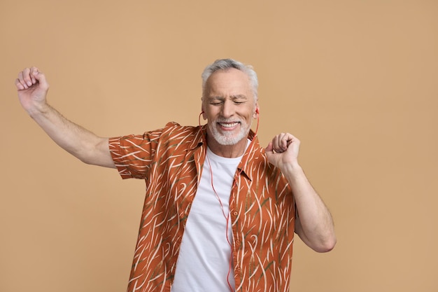 Un hombre mayor caucásico y elegante, feliz turista jubilado, usando auriculares naranjas y una camisa informal, sonriendo ampliamente moviéndose al ritmo de la música escuchando una banda sonora genial sobre un fondo beige aislado