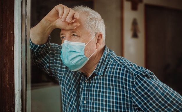 Hombre mayor en cuarentena por coronavirus, covid-2019 en una casa, mirando hacia afuera por la ventana