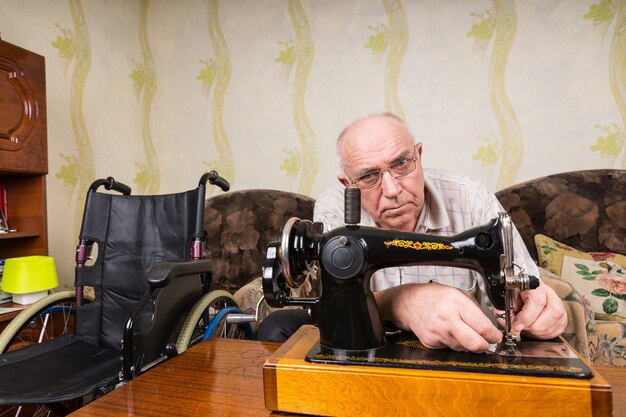 Hombre mayor con discapacidad usando anteojos mientras usa una máquina de coser mecánica vintage en casa, en una mesa de madera, cerca de un sofá y una silla de ruedas