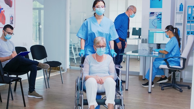 Hombre mayor discutiendo con la enfermera en la recepción del hospital con una máscara contra el coronavirus, el asistente con guantes estériles está empujando a una mujer madura discapacitada en silla de ruedas a través del área de espera.