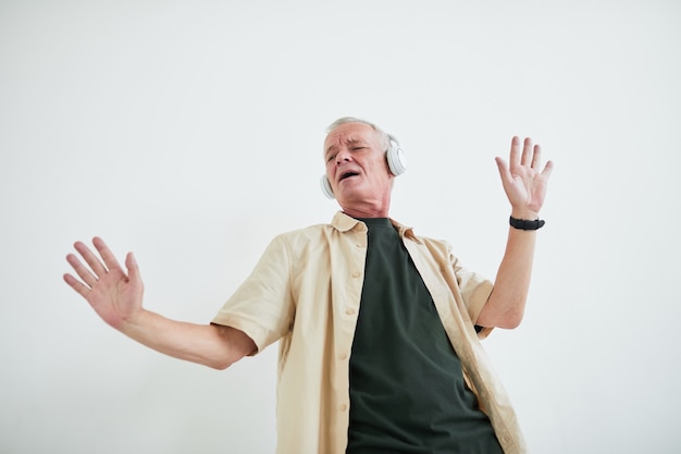 Hombre mayor emocionado escuchando música en auriculares inalámbricos y bailando contra el fondo blanco.