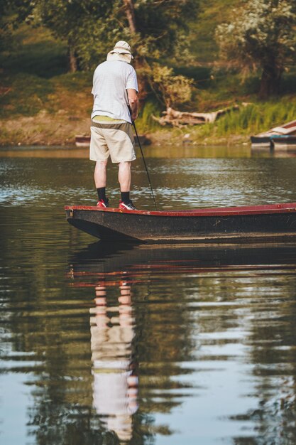 El hombre mayor está pescando desde un bote de madera en el centro de un lago de agua dulce bordeado por un paisaje exuberante.