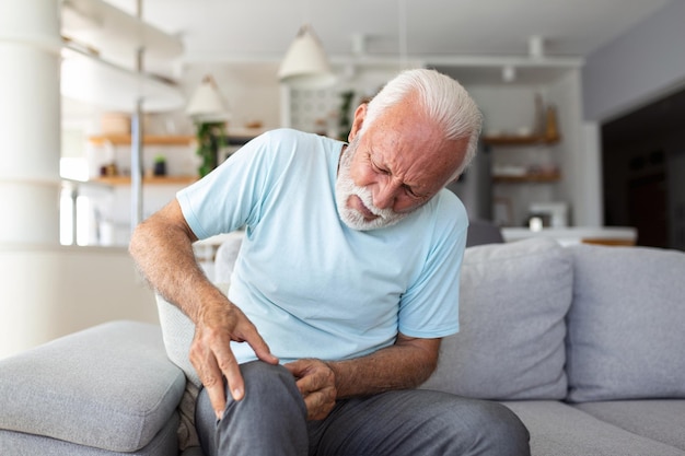 El hombre mayor está sentado en el sofá en casa tiene dolor en la articulación de la rodilla sosteniendo su pierna concepto de osteoartritis