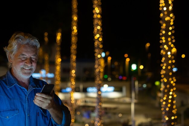 Hombre mayor feliz con barba blanca y cabello hablando por el teléfono celular. Al aire libre en la noche con palmeras iluminadas detrás de él. Decoraciones de luces amarillas.